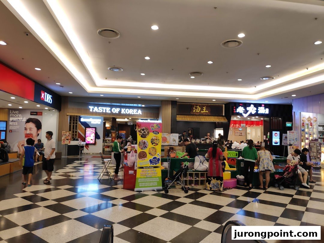 A bustling mall interior featuring various shop fronts, a promotional stand, and shoppers.