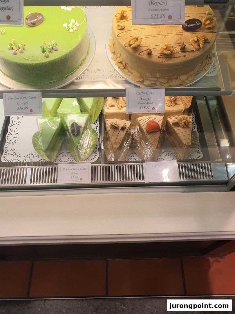 A display case showcasing a variety of delicious cakes, including Pandan Kaya and Coffee cakes, with visible price tags.