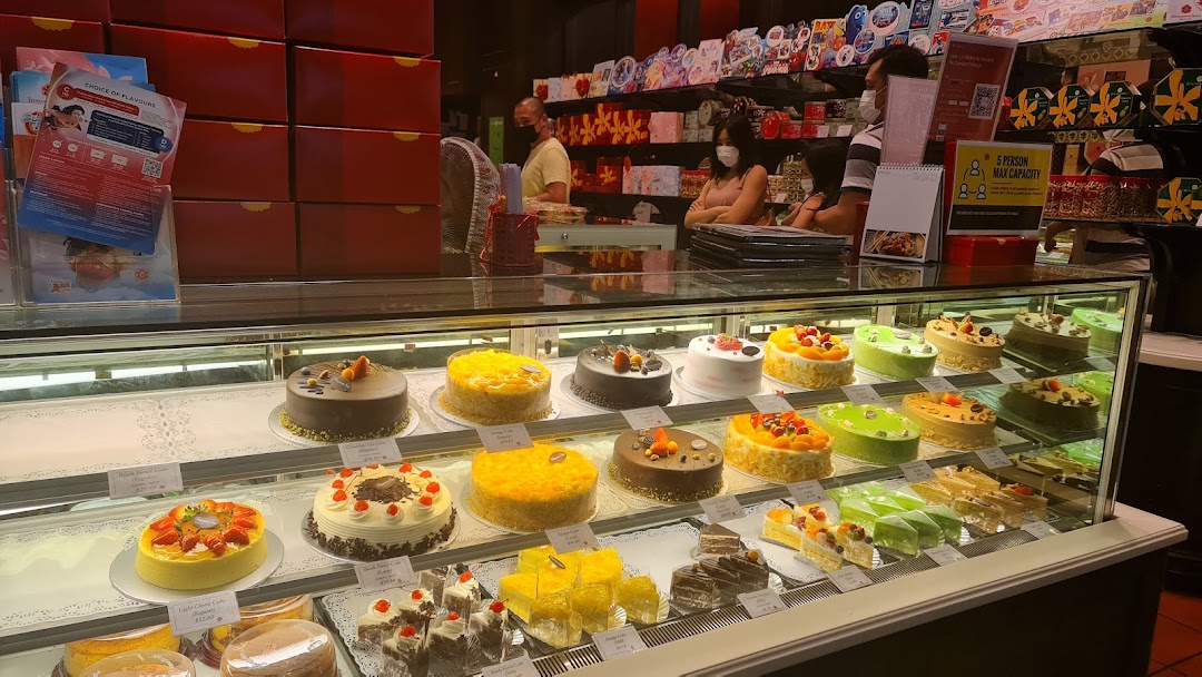 A vibrant display of various cakes and pastries in a well-lit bakery shop, with staff visible behind the counter.