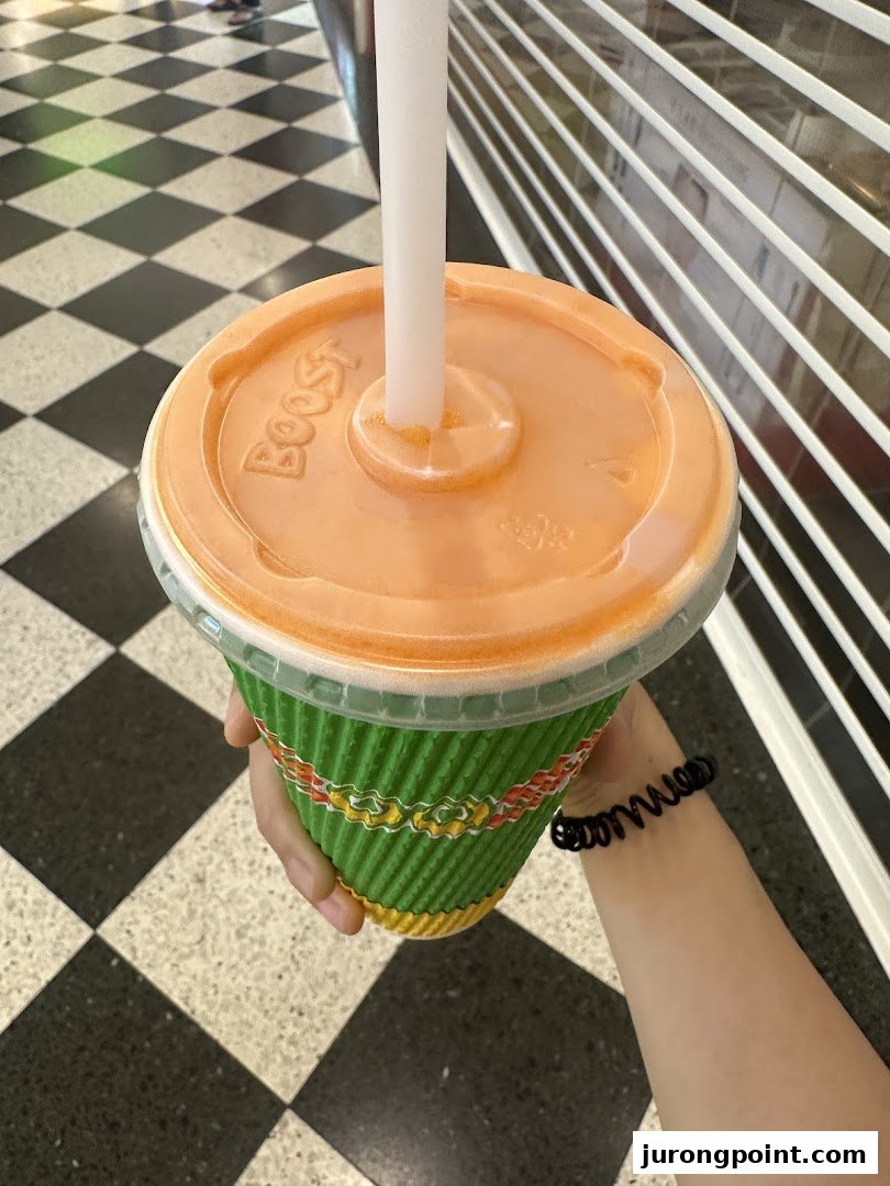 A hand holds a vibrant green Boost Juice cup with an orange lid and straw.