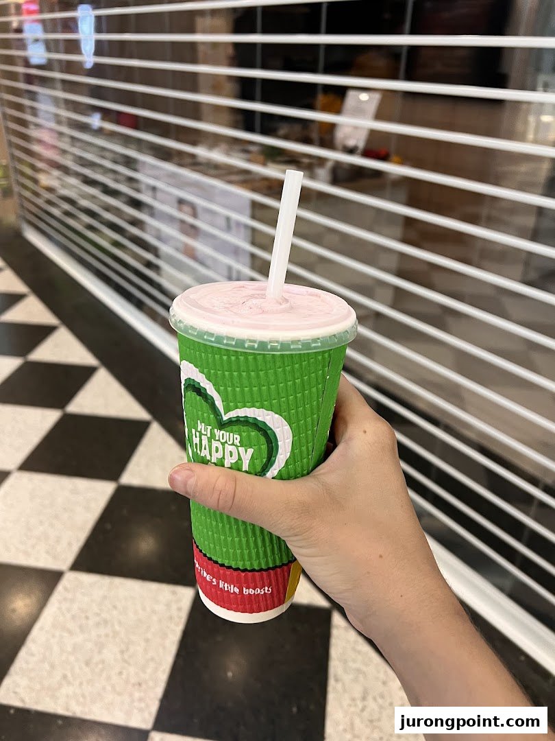 A hand holds a Boost Juice cup with a pink smoothie and straw, in front of a closed shop.