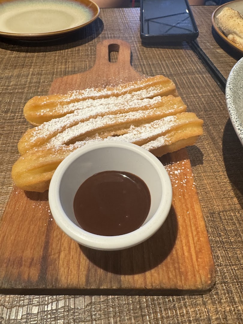 Golden churros dusted with powdered sugar, served with a rich chocolate dipping sauce on a wooden board.