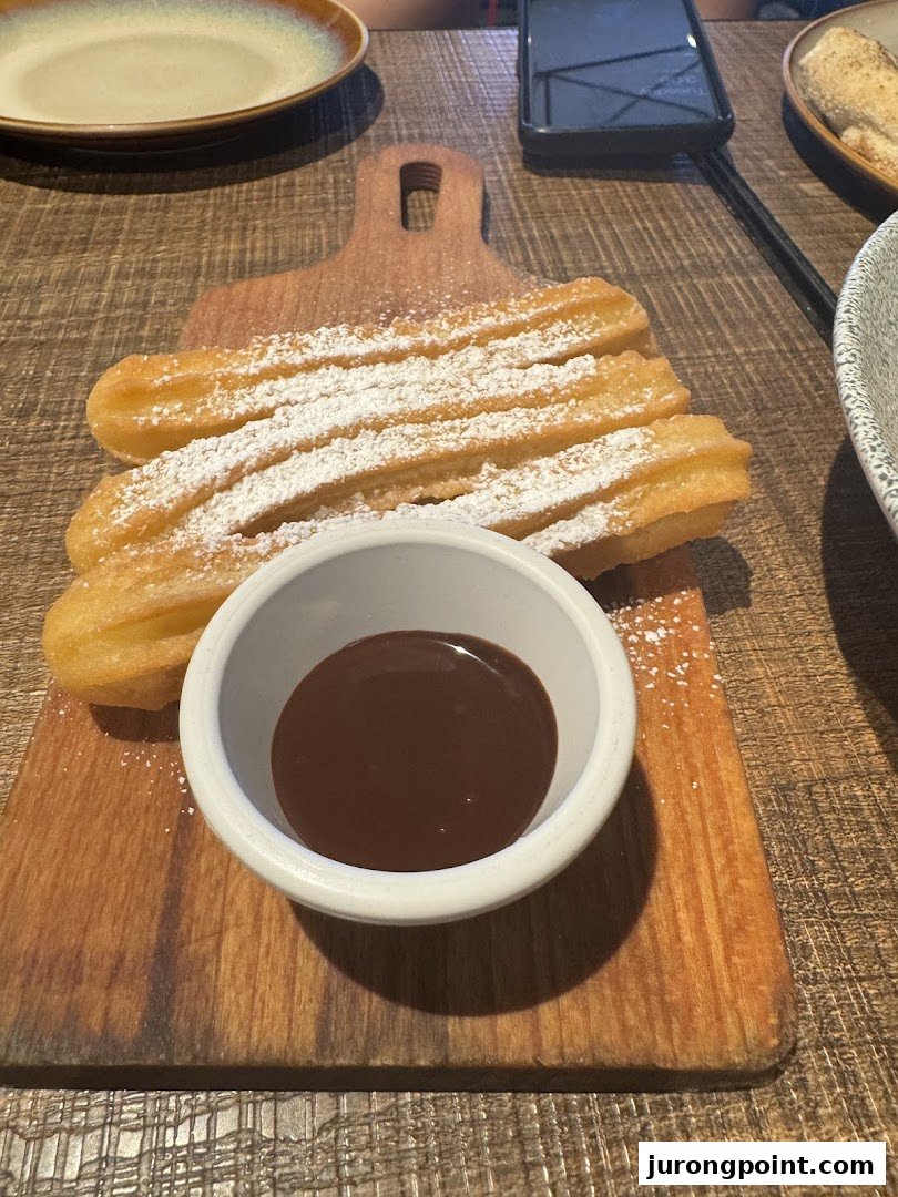 Golden churros dusted with powdered sugar, served with a rich chocolate dipping sauce on a wooden board.