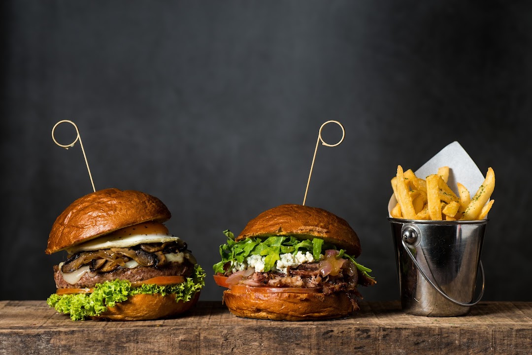 Two gourmet burgers with various toppings and a bucket of golden fries on a wooden surface.