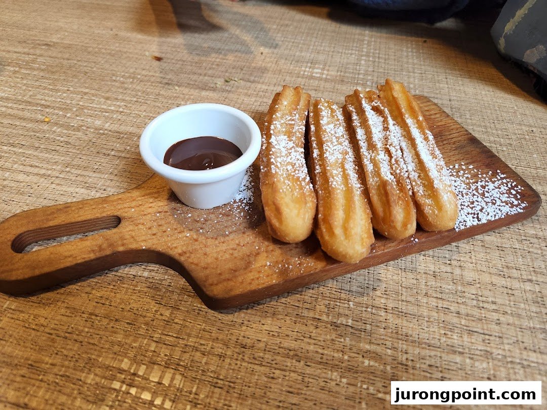 Five sugar-dusted churros and a ramekin of chocolate dipping sauce on a wooden board.