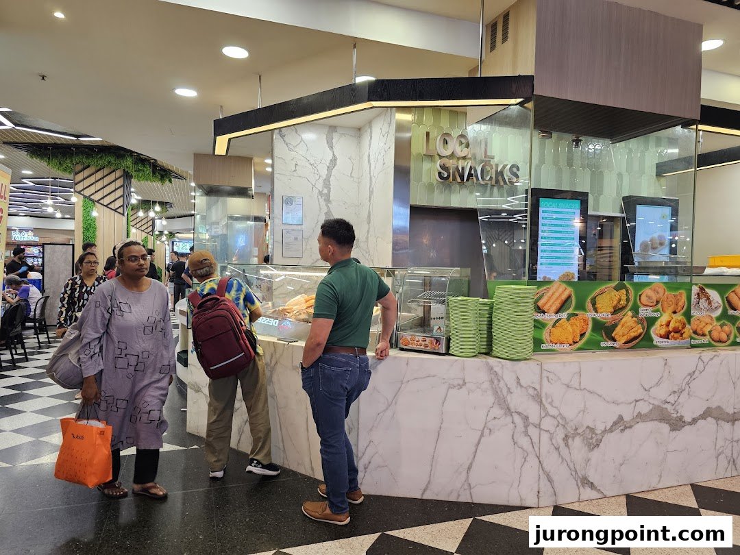 A bustling food stall counter featuring local snacks and a digital menu display within a mall.