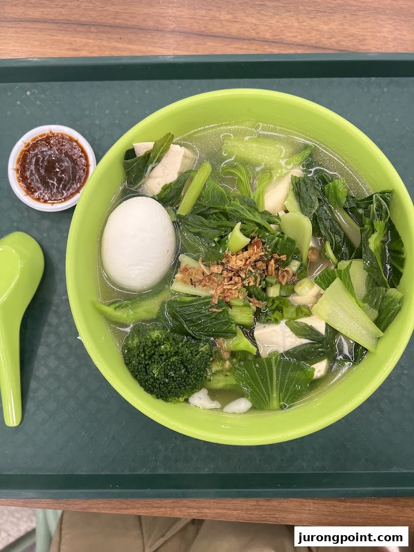 A vibrant bowl of clear soup with fresh green vegetables, tofu, and a hard-boiled egg, served with chili sauce.