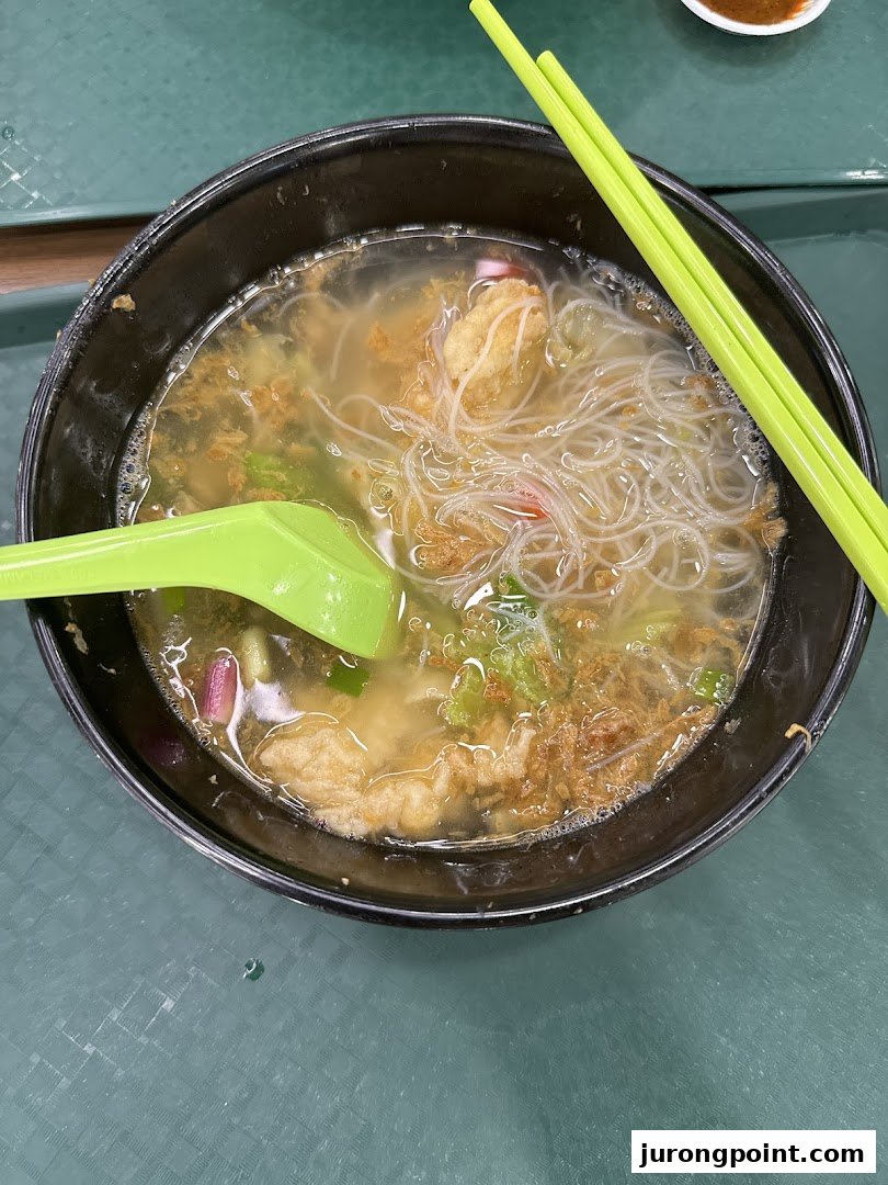 A close-up of a bowl of clear noodle soup with various ingredients and green chopsticks.