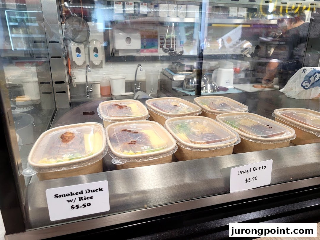Pre-packaged meals like Smoked Duck with Rice and Unagi Bento displayed with prices in a glass case.
