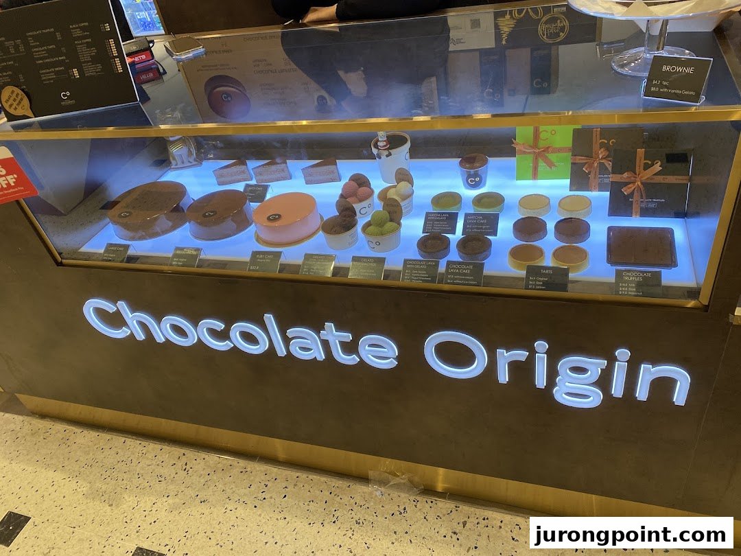 A brightly lit display case at Chocolate Origin showcasing a variety of decadent cakes, tarts, and chocolates.