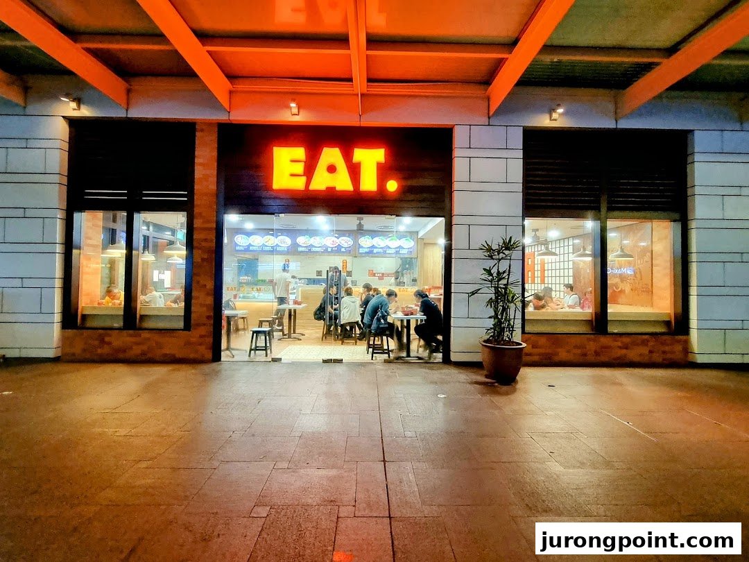 The brightly lit exterior of EAT. restaurant at night, with customers dining inside.