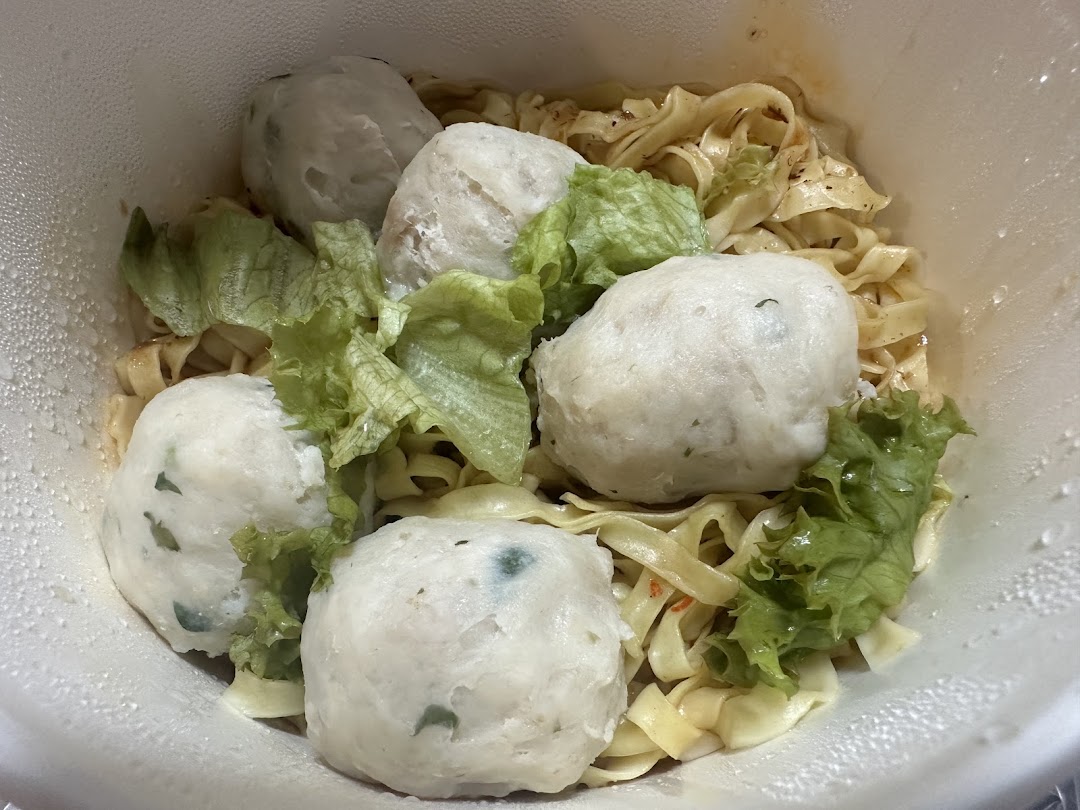 A close-up of noodles with large fish balls and lettuce in a white takeout container.