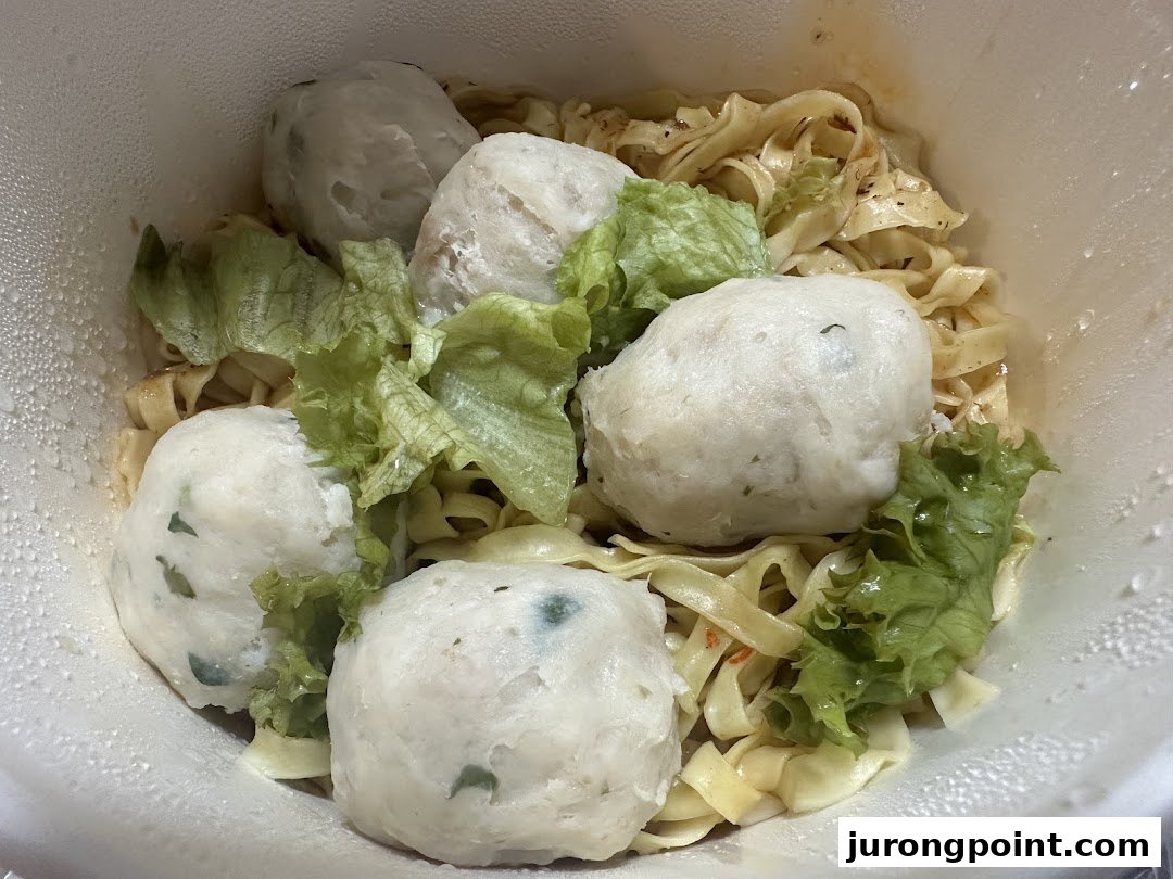 A close-up of noodles with large fish balls and lettuce in a white takeout container.