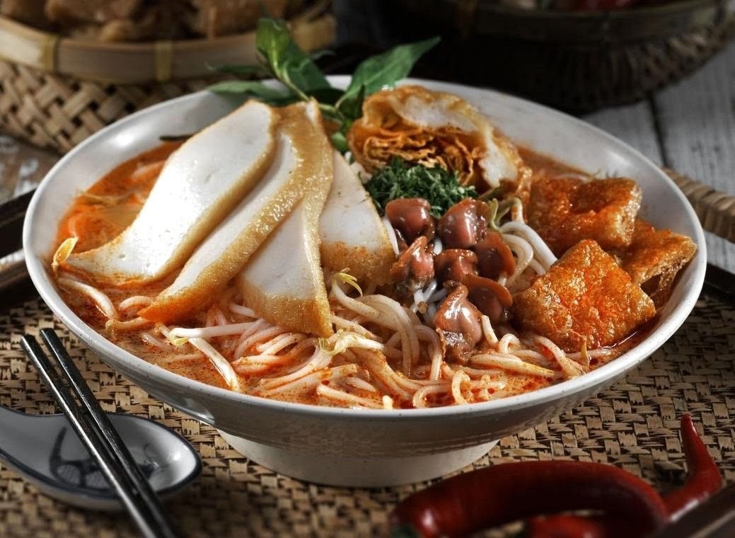 A vibrant bowl of spicy laksa noodles with fish cakes, cockles, and tofu puffs.