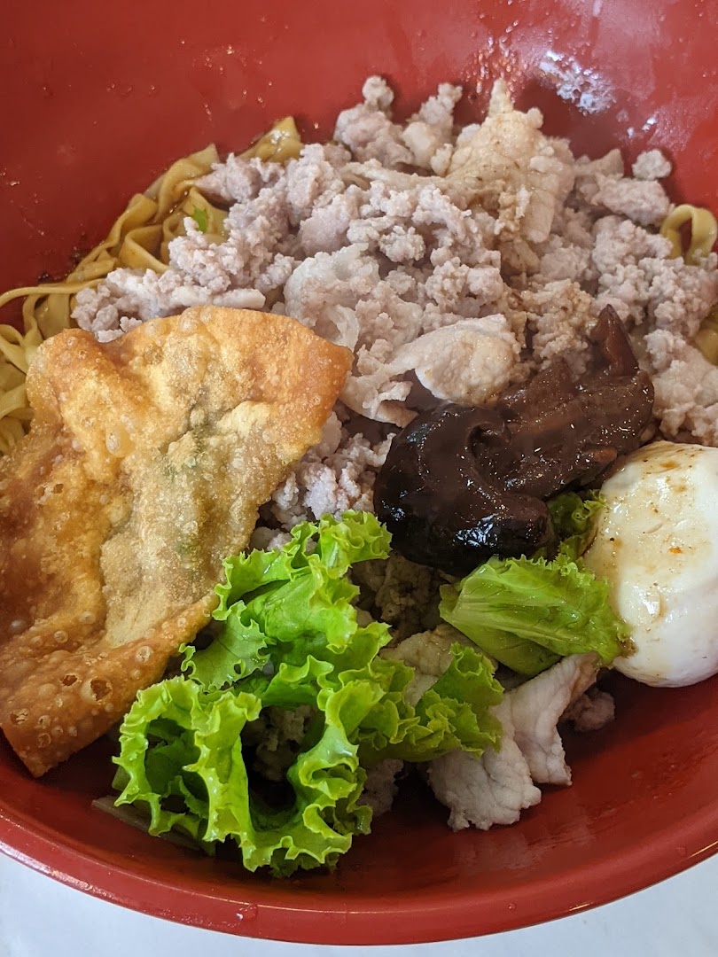A vibrant red bowl filled with noodles, minced meat, a fried wonton, fresh lettuce, and a dark sauce.