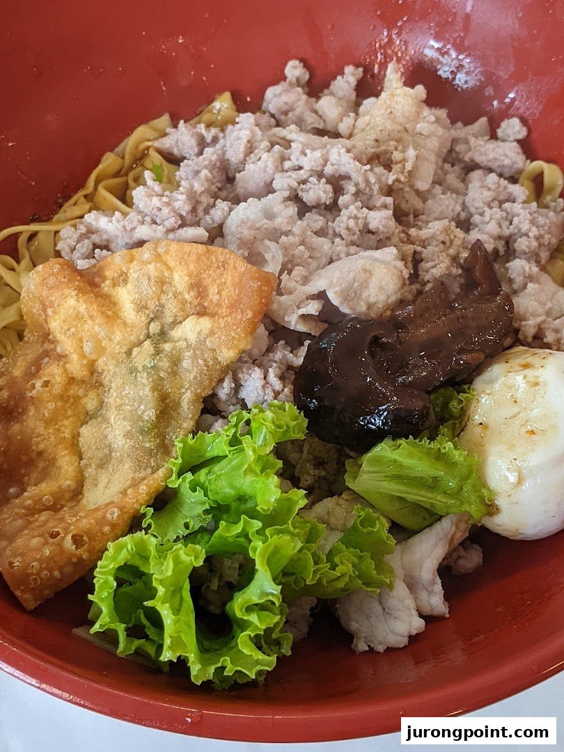A vibrant red bowl filled with noodles, minced meat, a fried wonton, fresh lettuce, and a dark sauce.
