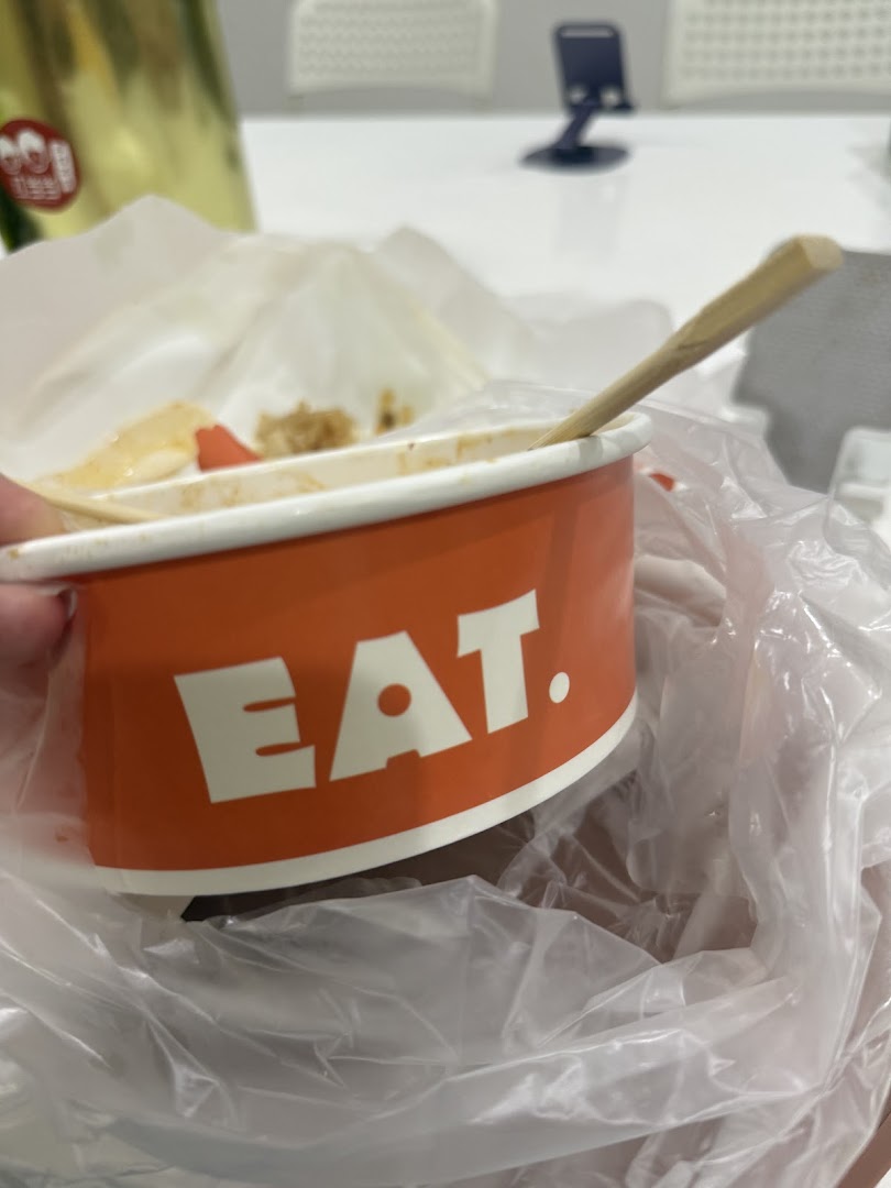 A partially eaten meal in an orange 'EAT.' branded container with a wooden spoon, inside a plastic bag.
