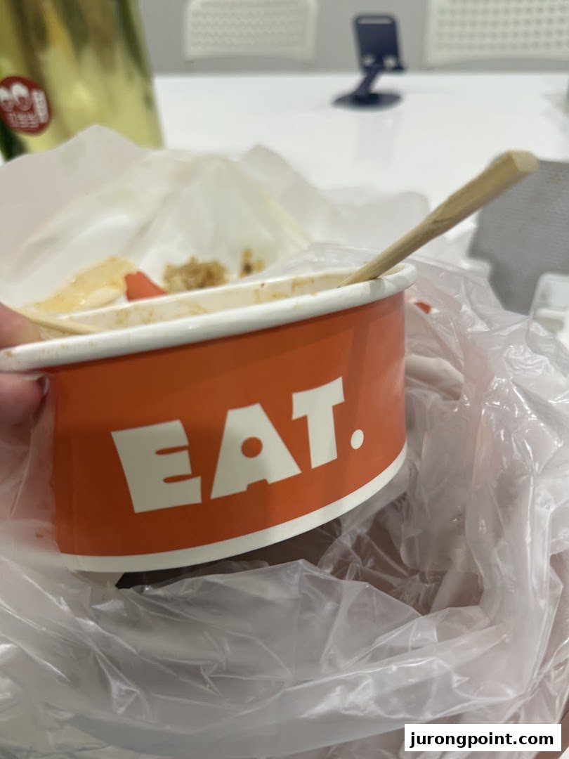 A partially eaten meal in an orange 'EAT.' branded container with a wooden spoon, inside a plastic bag.