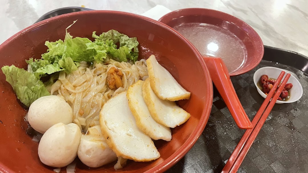 A close-up of a bowl of noodles with fish balls, fish cake slices, and lettuce, served with a side of soup and chili.