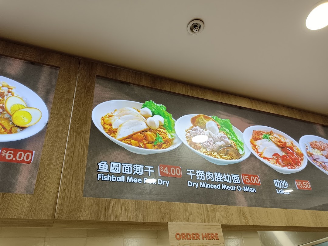 A brightly lit menu board displaying various noodle dishes with prices at a food stall.