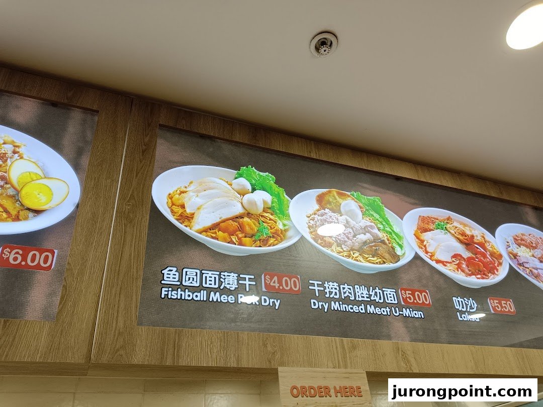 A brightly lit menu board displaying various noodle dishes with prices at a food stall.