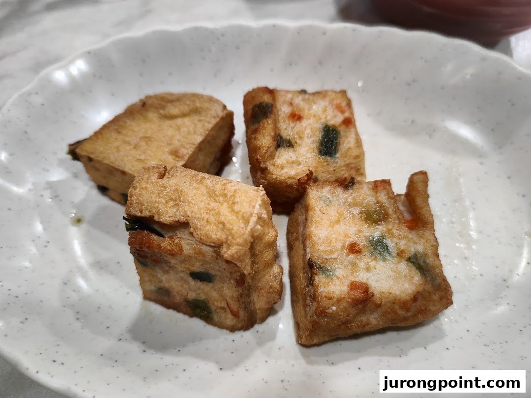 A close-up of four pieces of golden-fried, savory stuffed tofu or fish cakes on a white plate.