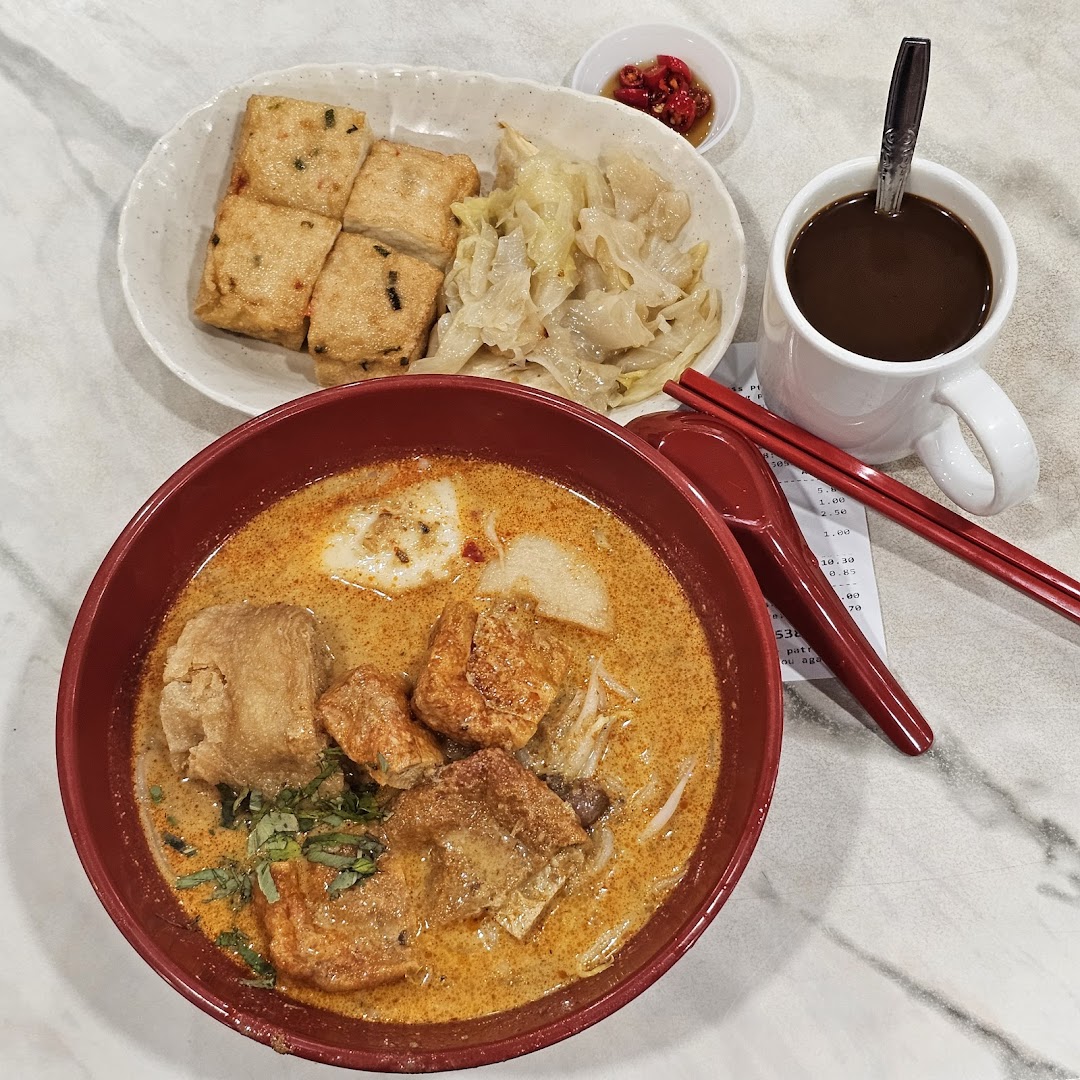 A vibrant bowl of curry noodles with tofu, braised cabbage, and a cup of coffee on a marble table.