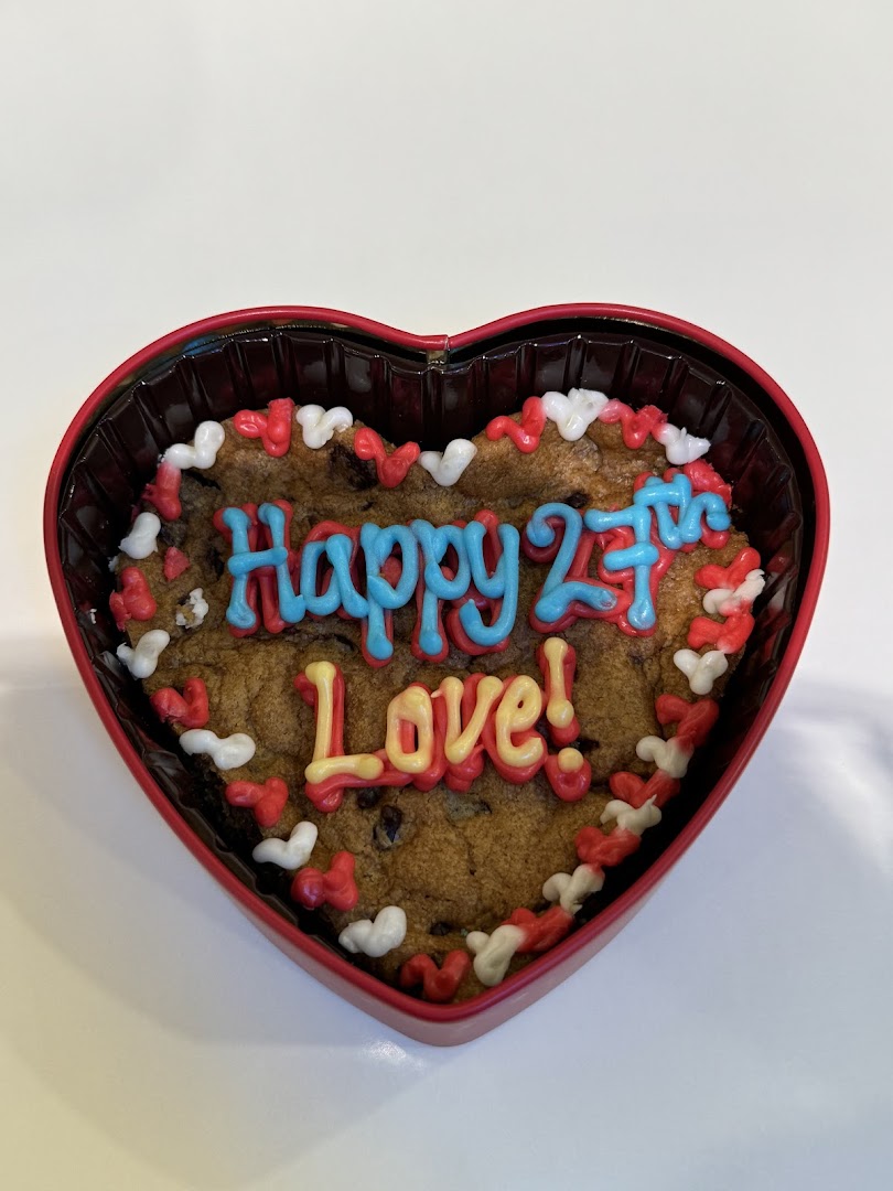 A heart-shaped chocolate chip cookie cake with colorful icing that reads "Happy 27th Love!" and sprinkles.