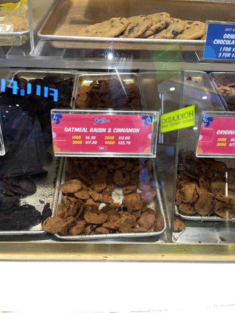 A display case at Famous Amos showcasing various cookies with visible pricing for Oatmeal Raisin & Cinnamon.