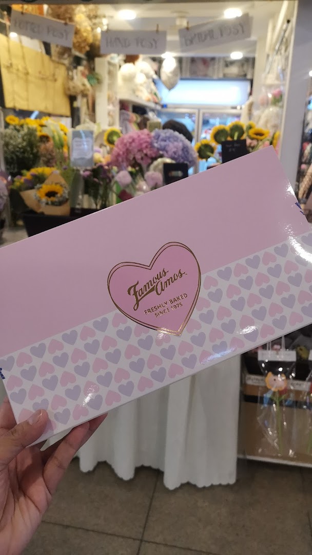 A hand holds a pink Famous Amos cookie box with heart patterns, featuring the brand logo.