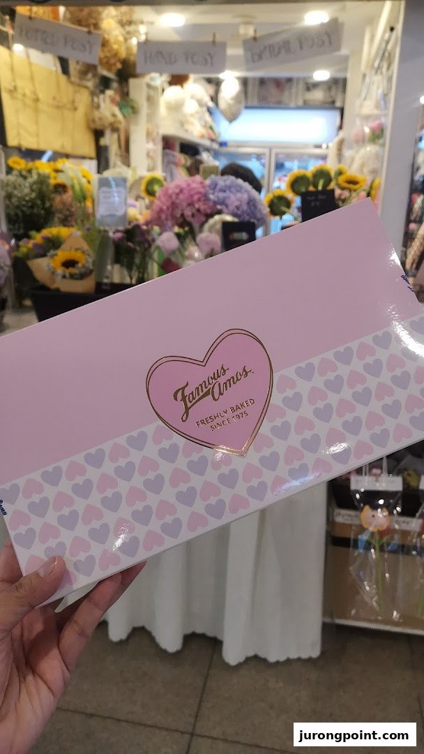 A hand holds a pink Famous Amos cookie box with heart patterns, featuring the brand logo.