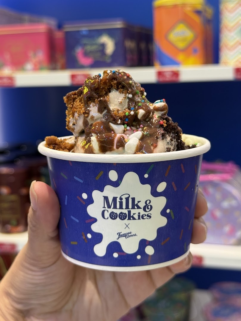 A hand holds a cup of 'Milk & Cookies x Famous Amos' dessert, topped with chocolate, sprinkles, and cookie pieces.
