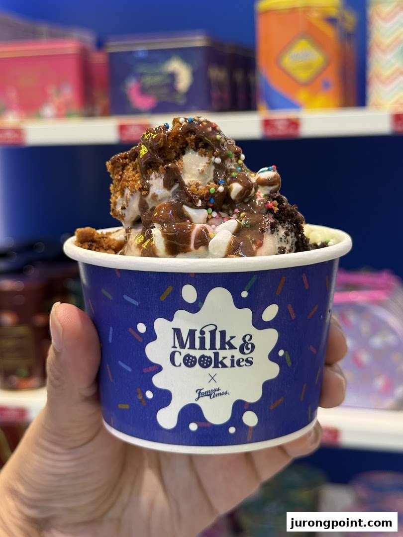 A hand holds a cup of 'Milk & Cookies x Famous Amos' dessert, topped with chocolate, sprinkles, and cookie pieces.