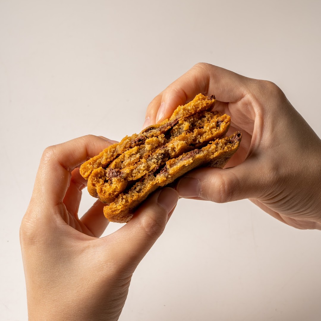 Hands gently break apart a warm, chewy Famous Amos chocolate chip cookie, revealing its gooey center.