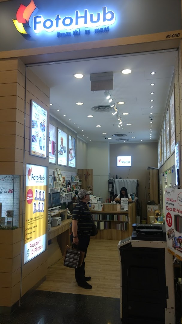 A customer stands inside FotoHub, a photo printing and ID photo service shop, with staff visible at the counter.