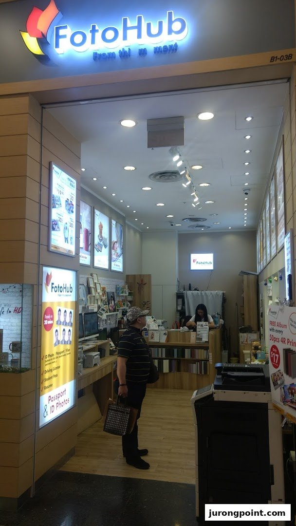 A customer stands inside FotoHub, a photo printing and ID photo service shop, with staff visible at the counter.