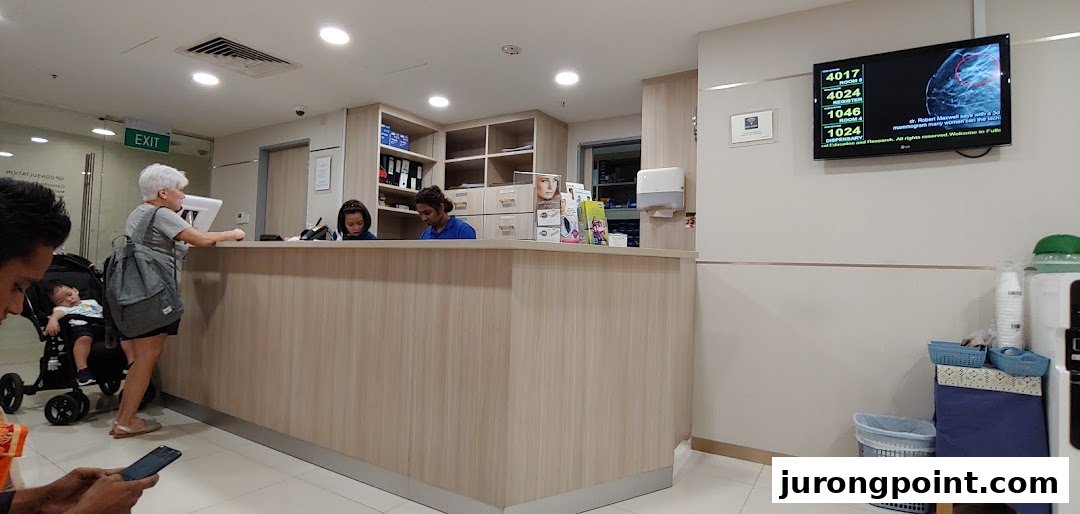 Fullerton Health reception area with staff assisting a patient, featuring a queue number display.
