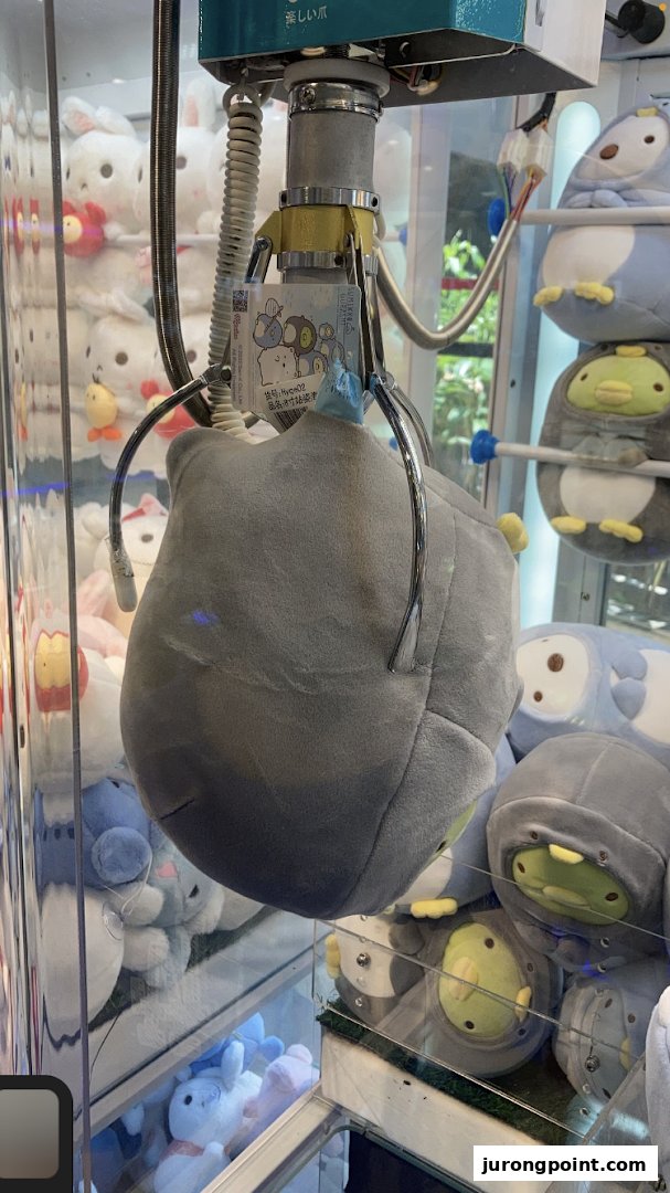 A claw machine's grabber holds a large plush toy, surrounded by other prizes inside the game.
