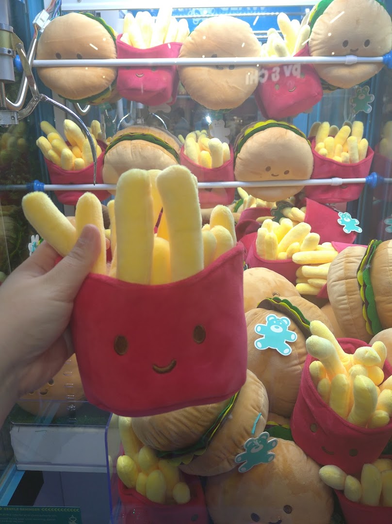 A hand holds a plush fries toy in front of a claw machine filled with burger and fries plushies.