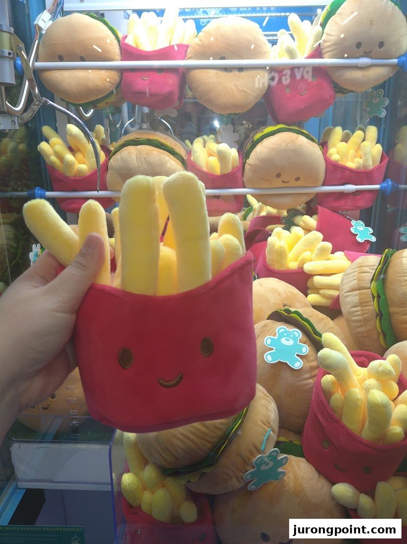 A hand holds a plush fries toy in front of a claw machine filled with burger and fries plushies.