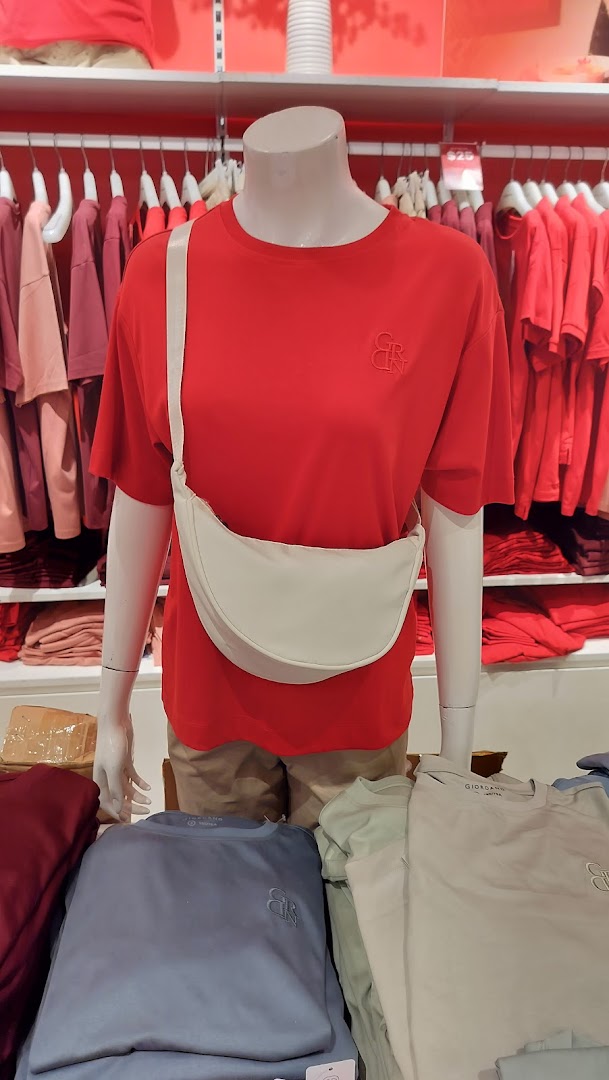 A mannequin displays a red t-shirt and white sling bag, surrounded by folded apparel in a retail store.
