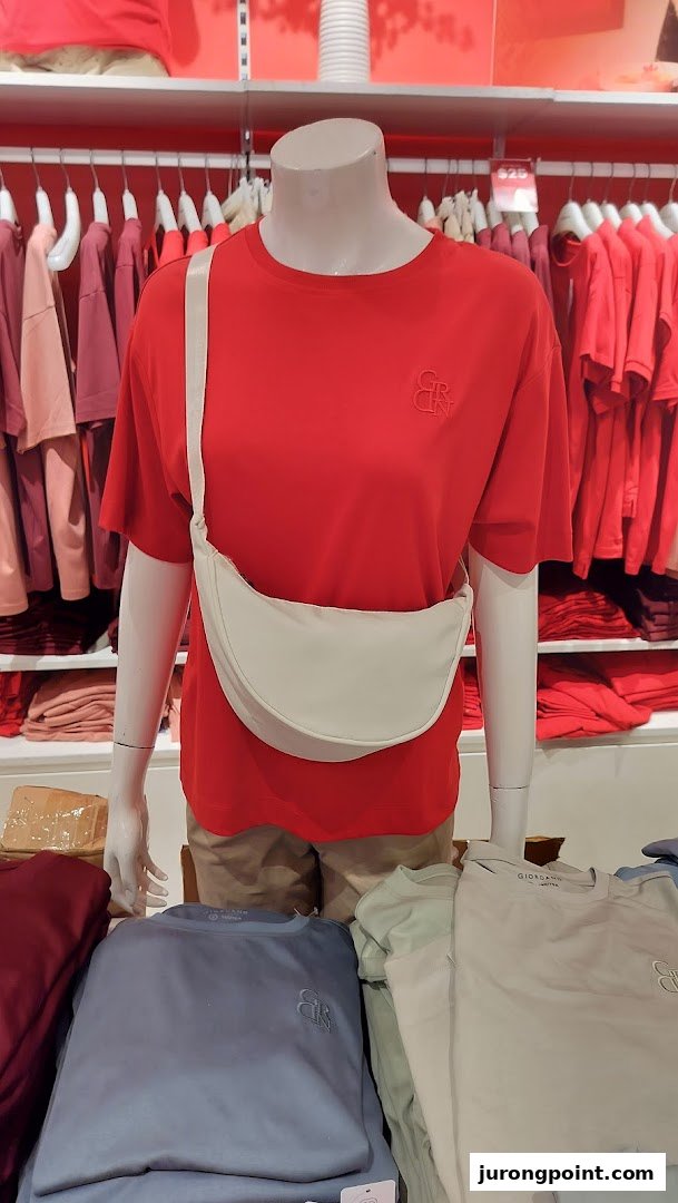 A mannequin displays a red t-shirt and white sling bag, surrounded by folded apparel in a retail store.