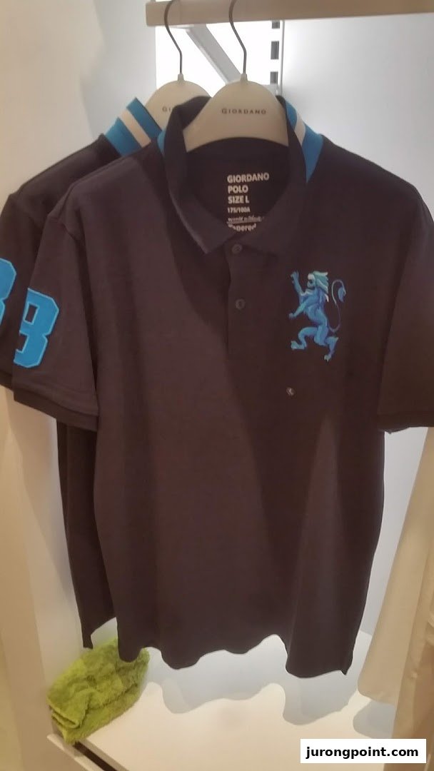 A dark Giordano polo shirt with a blue lion logo and striped collar hangs in a retail store.