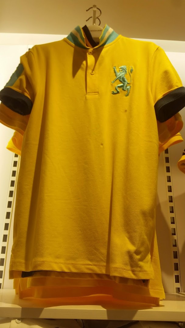 A vibrant yellow polo shirt with a green lion emblem and striped collar hangs in a retail store.