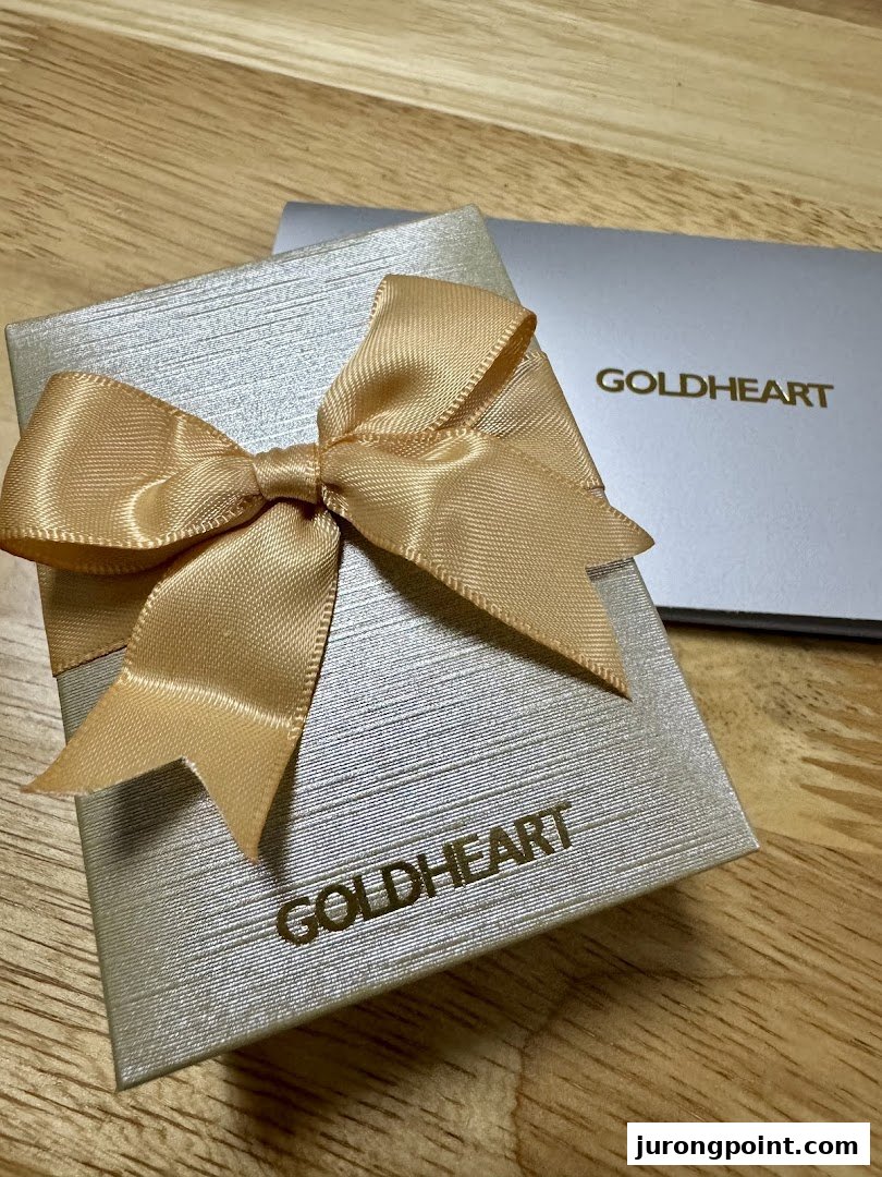 A silver Goldheart jewelry gift box with a gold ribbon bow on a wooden surface.