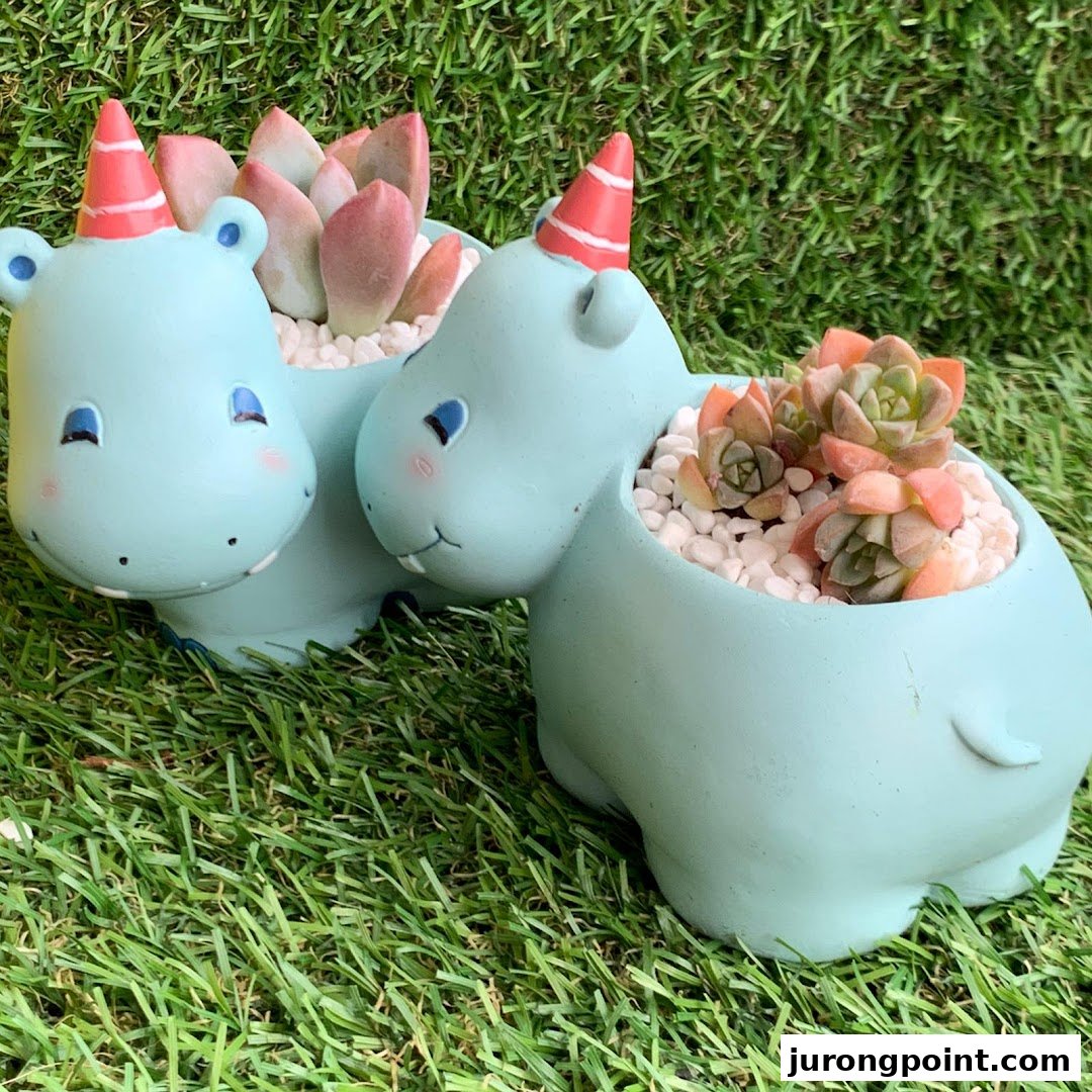 Two adorable light blue hippo planters with party hats, holding vibrant succulents on green grass.