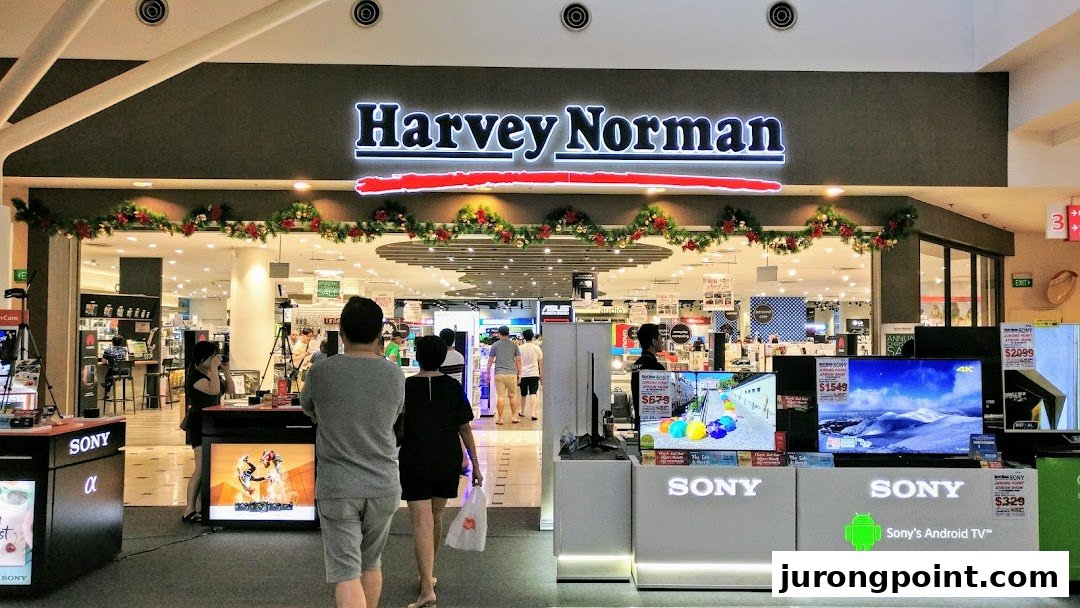 The entrance of Harvey Norman, an electronics and home goods store, featuring product displays and shoppers.