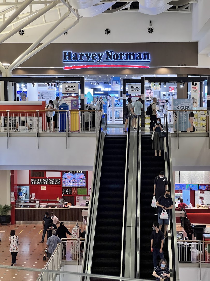 View of Harvey Norman shop entrance from a mall escalator, showcasing electronics and home goods.