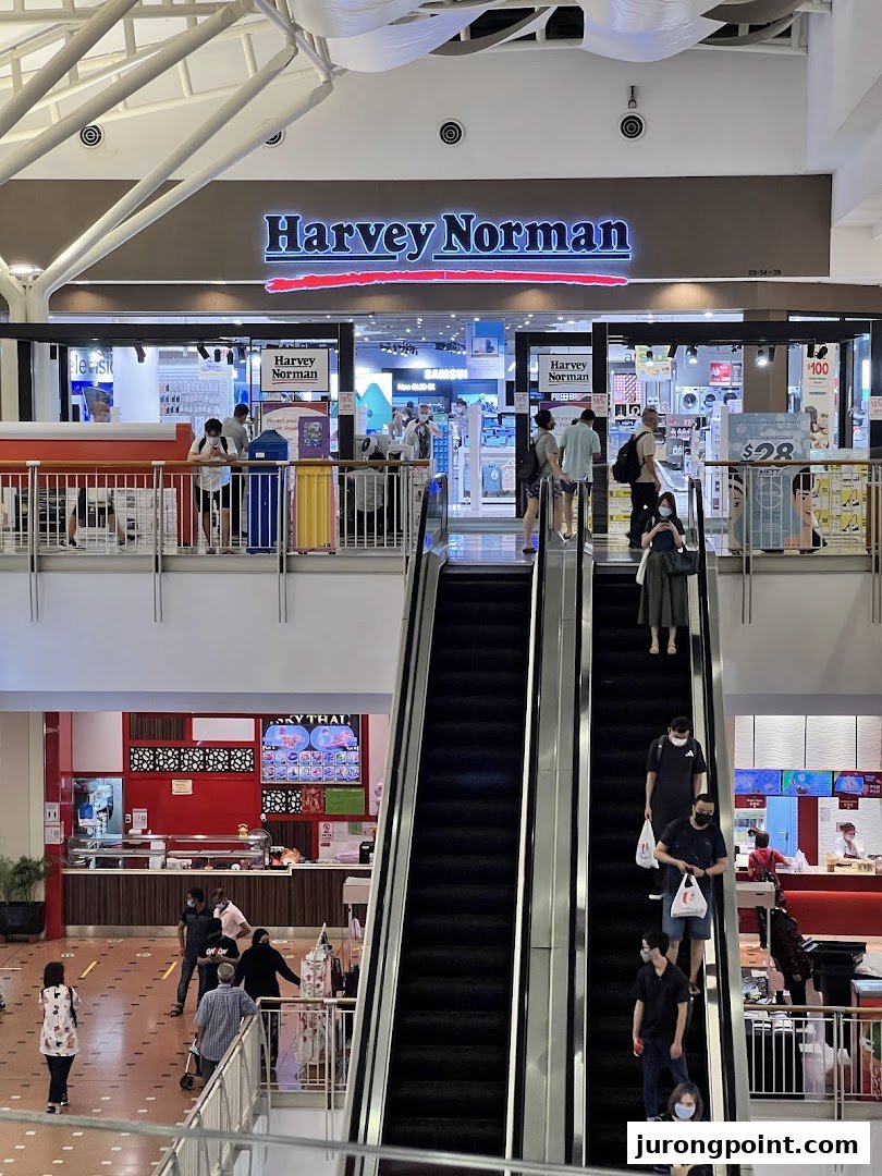 View of Harvey Norman shop entrance from a mall escalator, showcasing electronics and home goods.