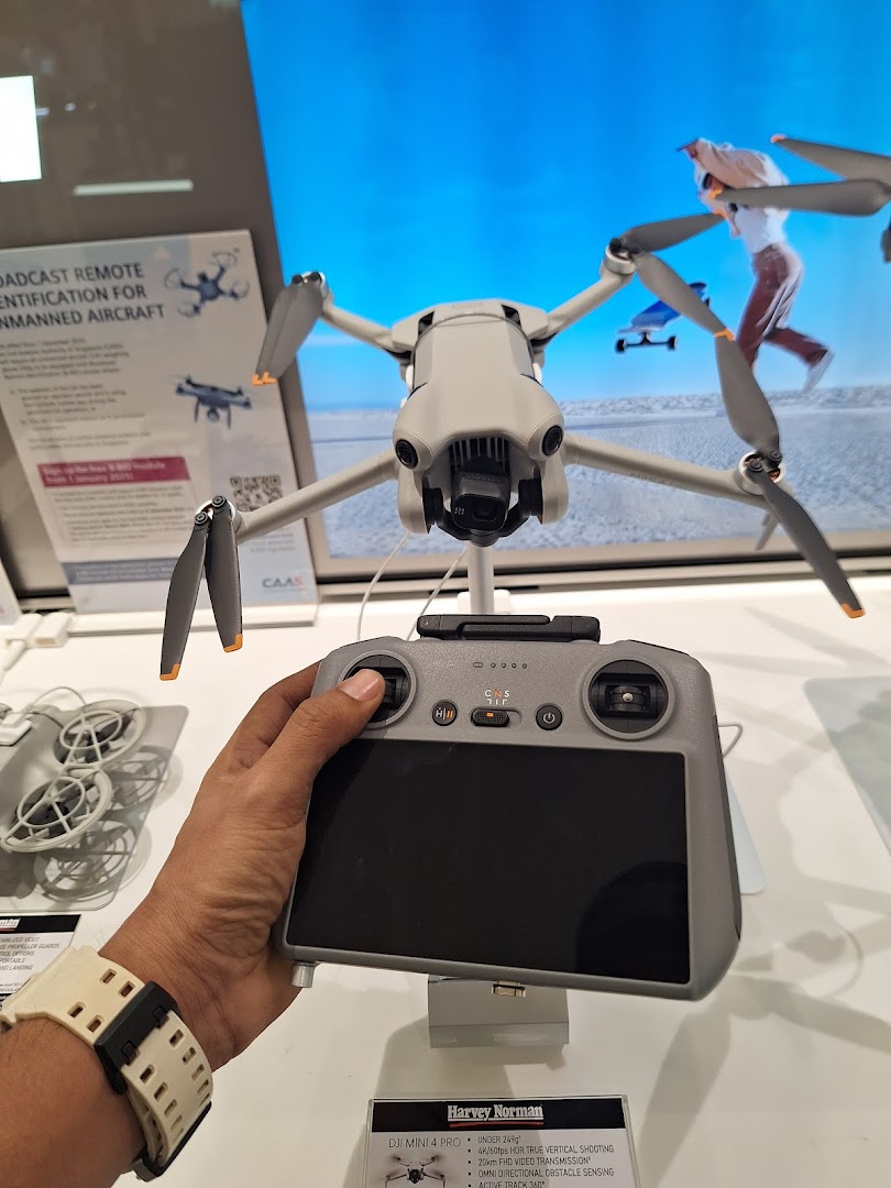 A customer's hand holds a DJI drone controller, with a DJI Mini 4 Pro drone on display at Harvey Norman.